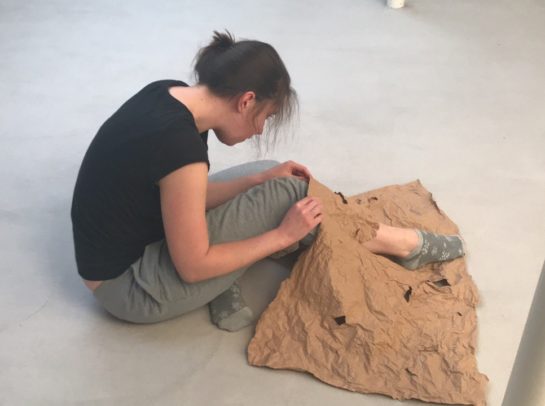 Butoh tanzende Person: Person sitzt am Boden, vor sich und teilweise auf den Beinen liegt ein zerknittertes Stück Papier mit einem Loch. Durch dieses Loch steckt sie einen Fuß. Der Blick ist auf den Fuß gerichtet.