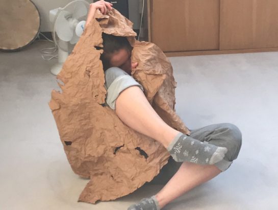 Butoh tanzende Person: am Boden sitzend mit einem zerknitterten Stück Papier. Sie hält das Papier vor sich. Im Papier ist ein Loch, durch das sie ein Bein Streckt.